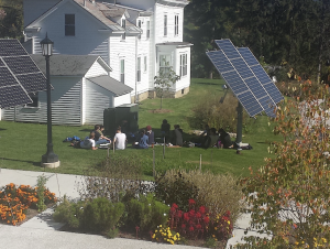 A class sitting on the grass behind one of the two solar trackers, half the students sitting in the shade and half sitting in the sun.